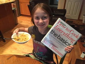 Little girl reading a cookbook and eating a bowl of pasta